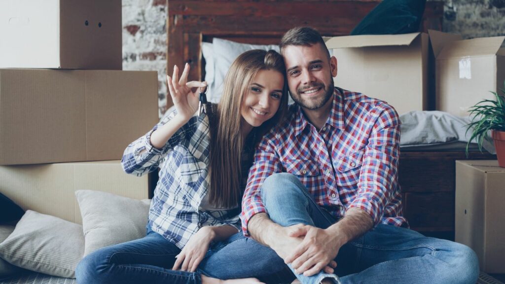 Smiling couple in new home holding keys, surrounded by moving boxes.