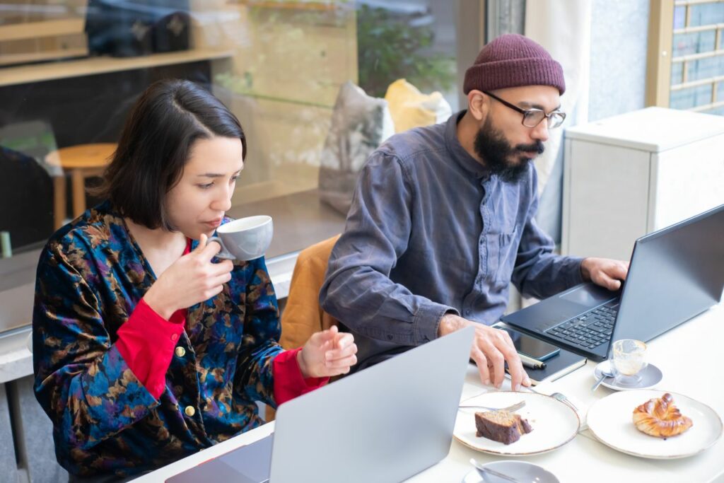 Two adults working on laptops in a contemporary café, enjoying coffee and pastries.